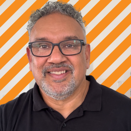 Mark Yettica-Paulson This is an image of Mark Yettica-Paulson. He has grey hair and a grey beard and moustache. He is wearing black frame glasses and a black polo shirt. There is a stripe orange and white background behind him.